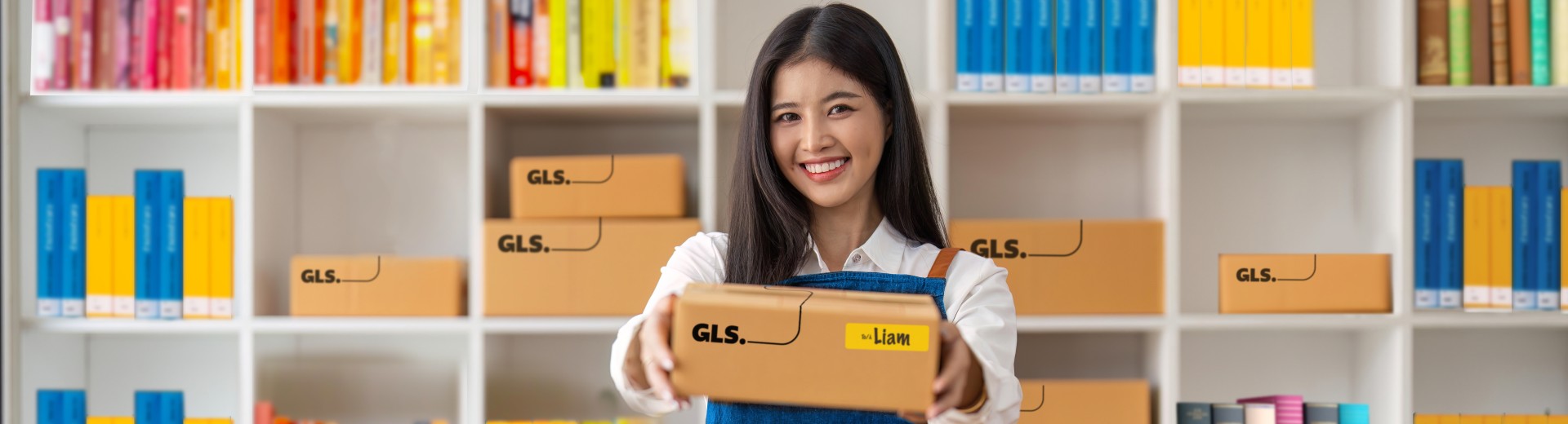 Woman handing out a GLS parcel. She stands at a desk in front of shelves field with colourful books. Woman handing out a GLS parcel. She stands at a desk in front of shelves field with colourful books.