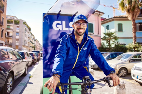Livreur GLS souriant sur un vélo-cargo électrique avec caisson bleu GLS, dans une rue urbaine ensoleillée.