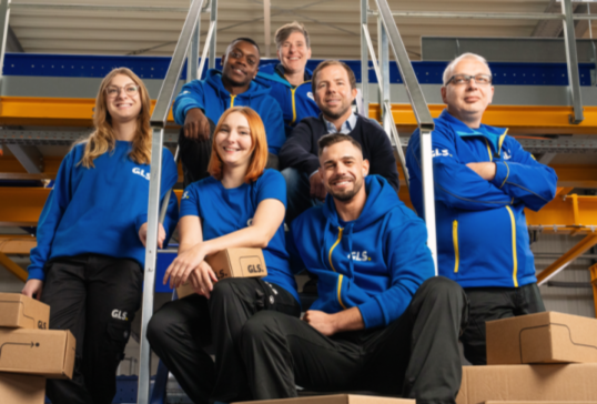 Groupe de collaborateurs GLS souriants et diversifiés, saluant la caméra dans un hall de bureaux moderne.