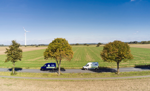 Deux camionnettes de livraison GLS sur une route de campagne, avec champs verts et éoliennes à l'horizon.