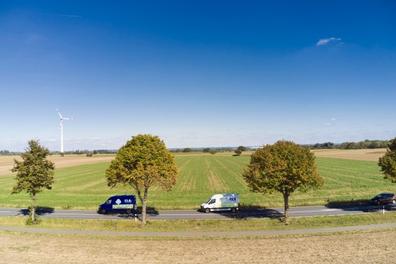 Deux camionnettes de livraison GLS roulent sur une route de campagne bordée d'arbres, au milieu de vastes champs cultivés. Une éolienne se dresse à l'horizon sous un ciel bleu dégagé, tandis qu'une voiture circule plus loin sur la même route.